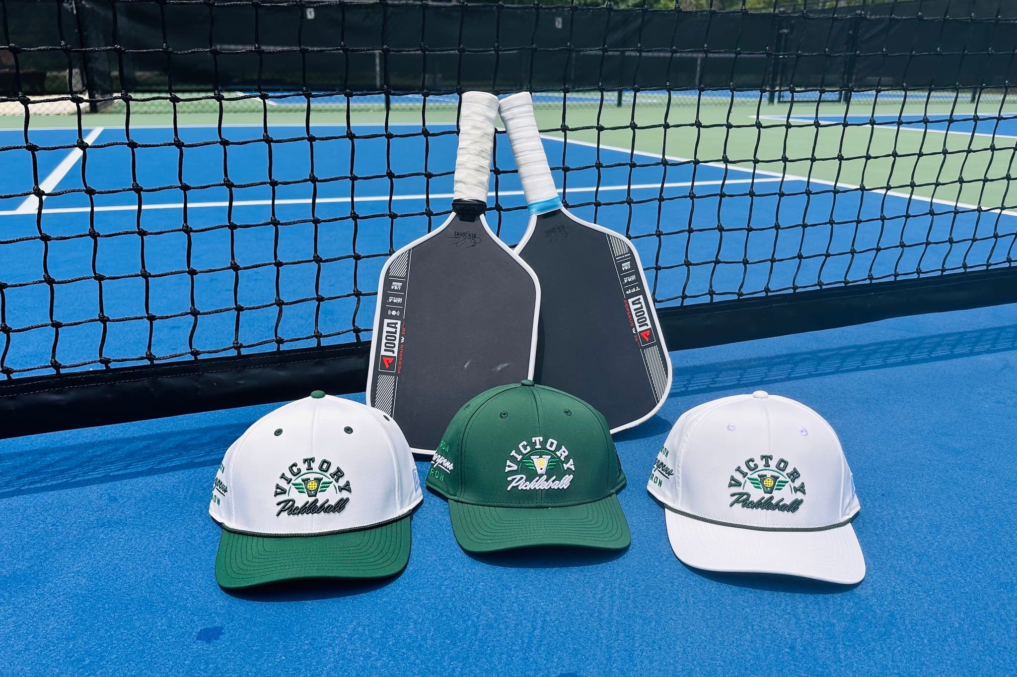 Three Victory Pickleball Academy caps and two paddles resting against the net on an outdoor court.
