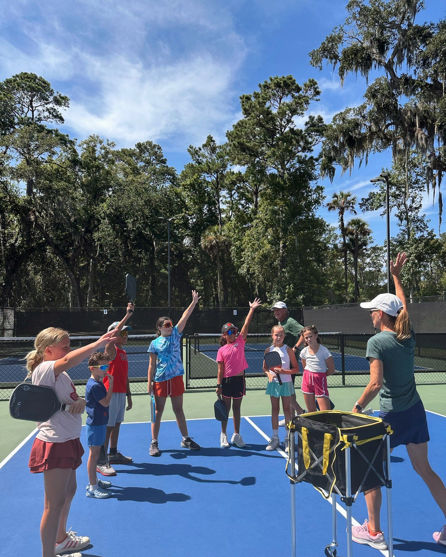 A group of students and two coaches raise their paddles overhead together at the end of a session, under a bright blue sky.