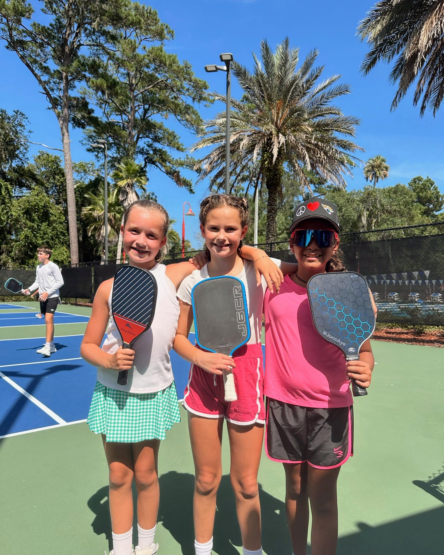 Three young players stand shoulder to shoulder with their arms around each other, holding their paddles up and smiling at the camera.