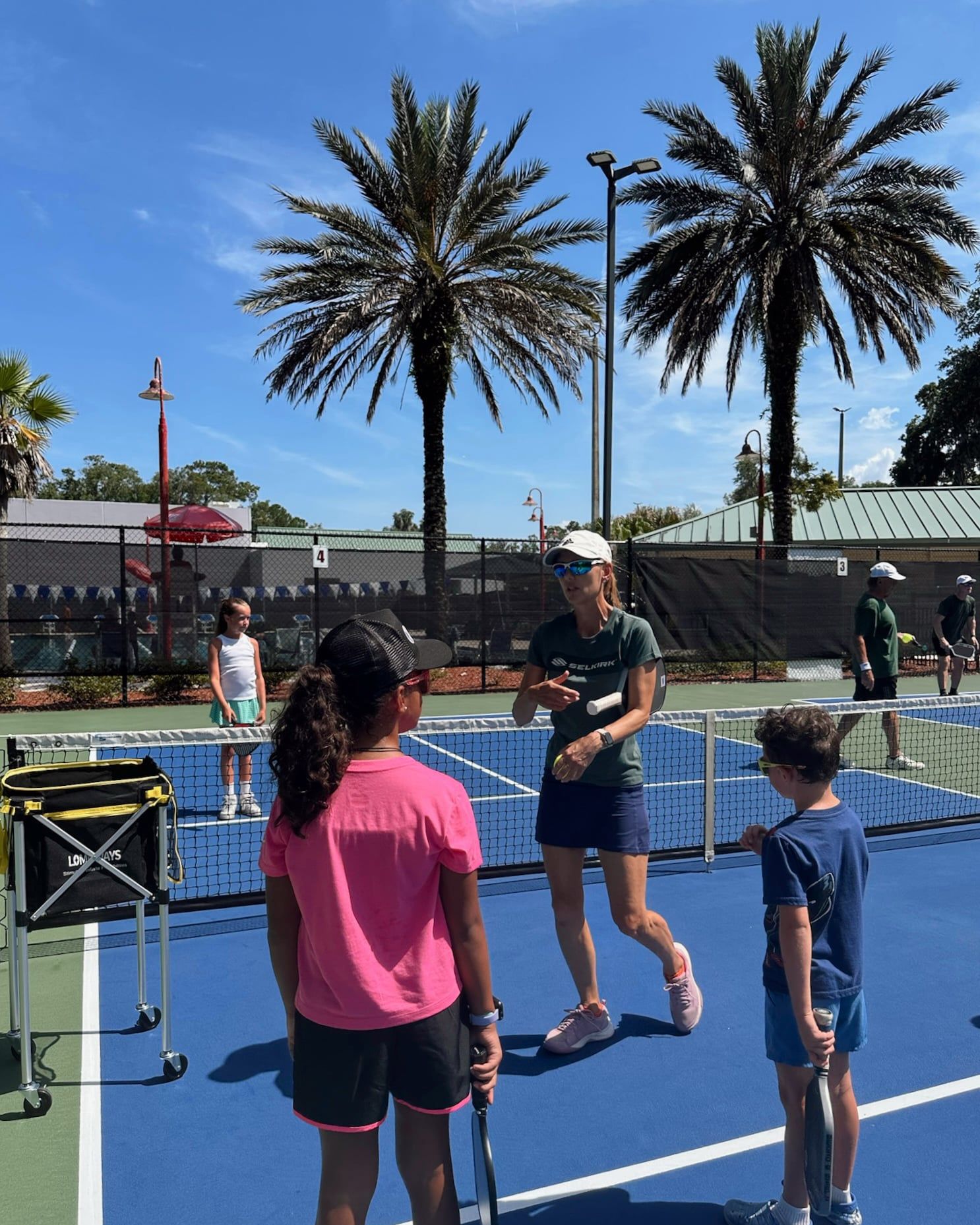 A coach demonstrates a paddle grip to two students beside a ball cart at the edge of the court.