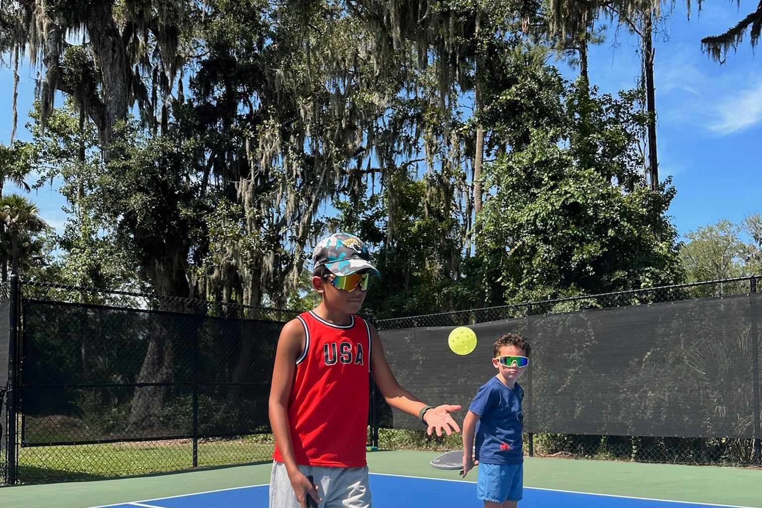 An older student tosses a ball underhand to a much younger player, who sets up to hit it back.