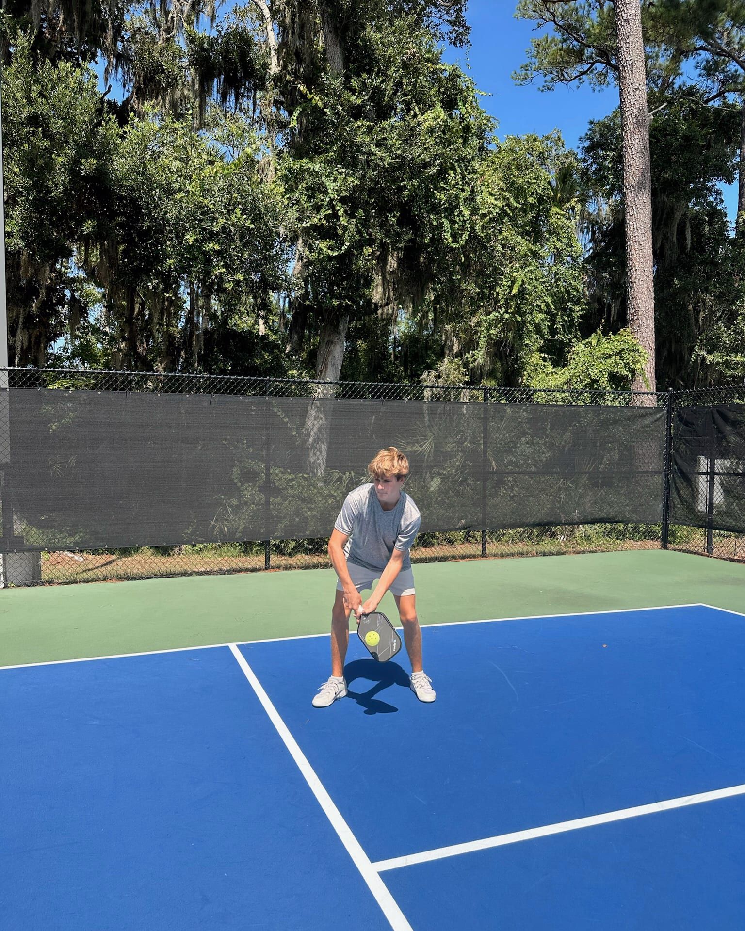 A young player crouches in a ready stance at the baseline of a blue outdoor pickleball court, paddle held in both hands, waiting to return serve.