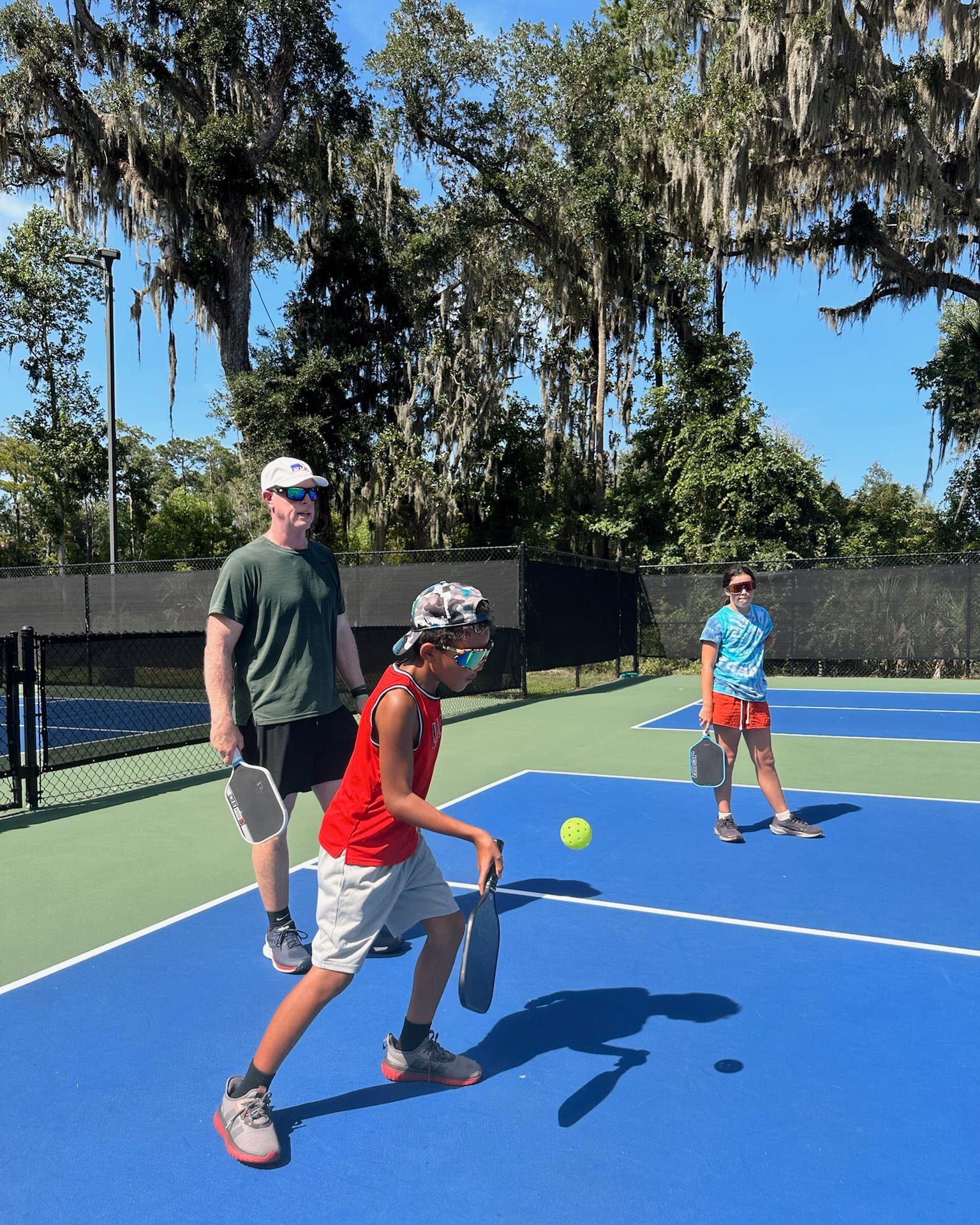 A young player strides forward to return the ball as two coaches watch from the far side of the court.