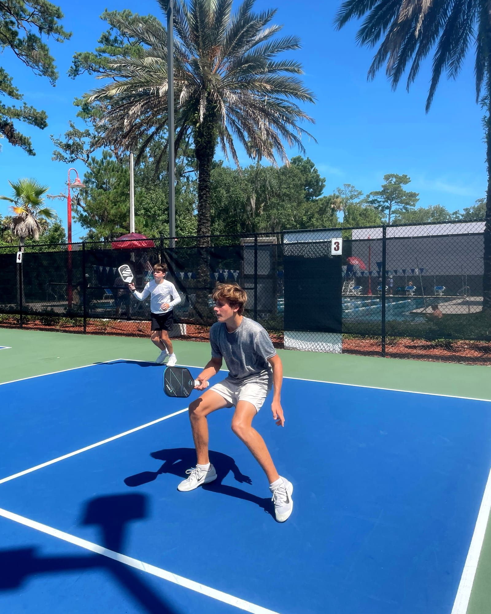 A wide view across two outdoor pickleball courts with a coach and around eight students spread out in play, the net crossing the foreground.