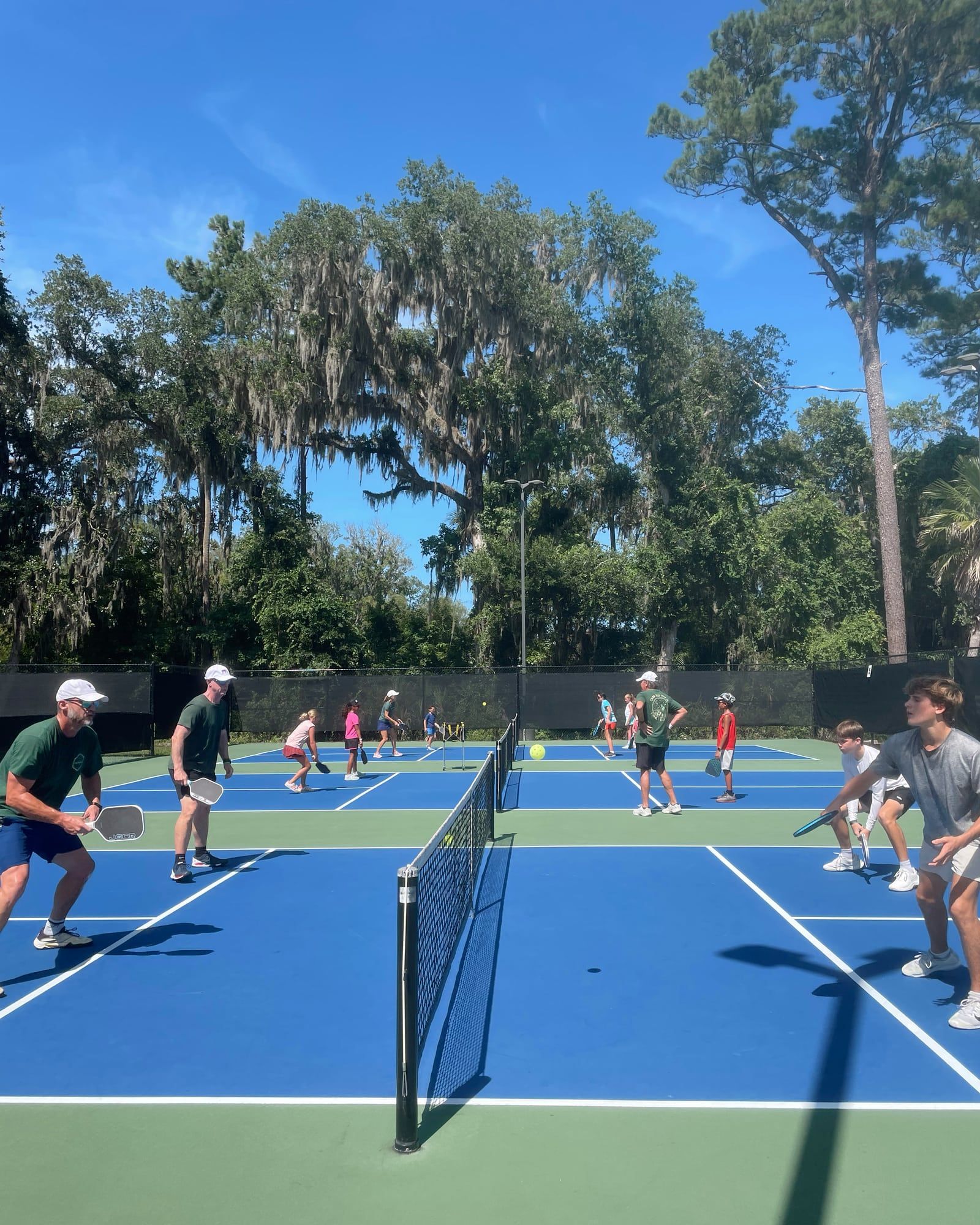 Two adjoining courts seen from behind the net, with coaches and students playing on both sides beneath tall pines.
